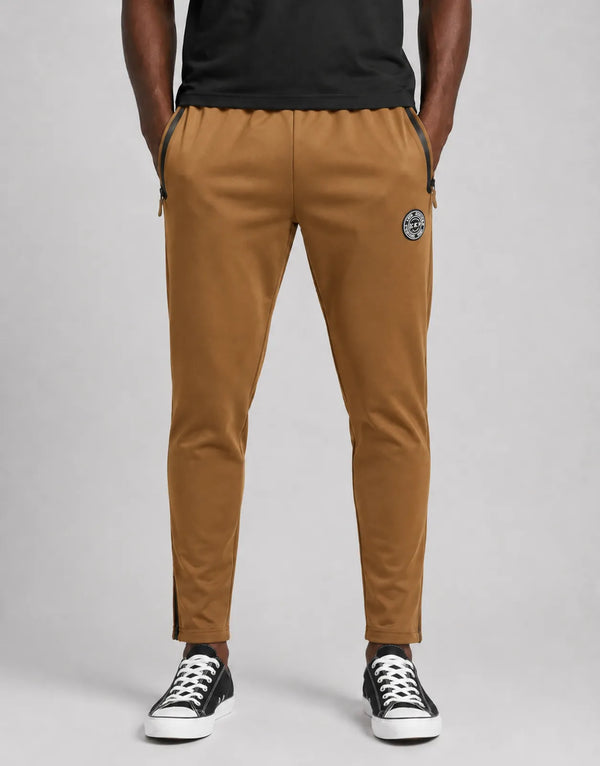 tan trackpants with black zip pockets and a round logo patch on the left thigh, shown with black and white sneakers