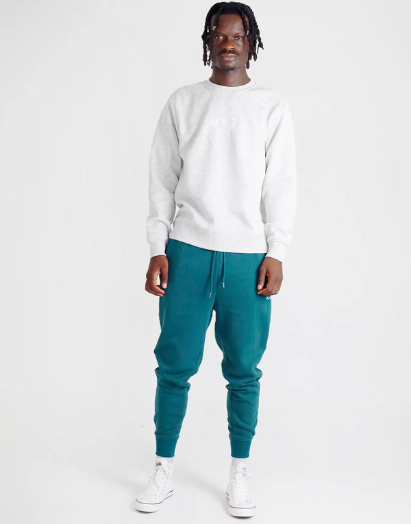 man wearing a light grey crew neck sweatshirt and teal jogger pants with drawstring and ribbed cuffs, white sneakers and a